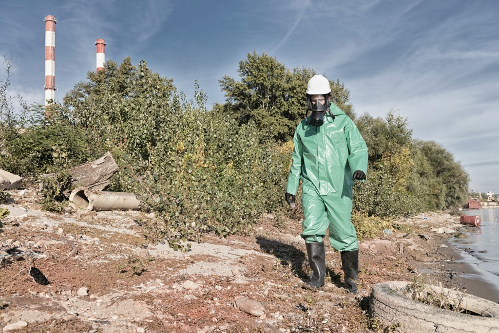 Environmentalist in protective suit working at a pollution site. Contamination of air, water and soil. Pollution factors like factory chimneys, old tires, plastic bottles, chemicals and other waste, all represented in the photo.